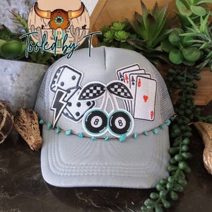 May include: A gray trucker hat with a mesh back. The hat has a variety of embroidered patches including dice, a lightning bolt, a cherry, a checkered flag, and playing cards. The hat is decorated with turquoise beads around the brim.