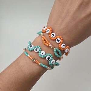 May include: Four beaded bracelets spelling out "SHOWGIRL" in white letter beads. The bracelets are in shades of orange, teal, and gold. The bracelets are worn on a wrist.