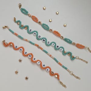 May include: Four beaded bracelets with the word "SHOWGIRL" spelled out in white letter beads. The bracelets feature teal, orange, and gold beads, with gold star charms. The bracelets are on a white surface with scattered gold star-shaped confetti.