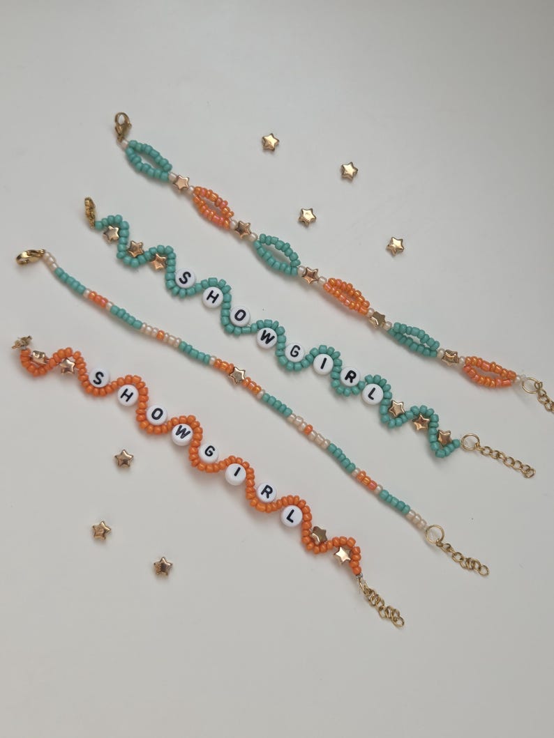 May include: Four beaded bracelets with the word "SHOWGIRL" spelled out in white letter beads. The bracelets feature teal, orange, and gold beads, with gold star charms. The bracelets are on a white surface with scattered gold star-shaped confetti.