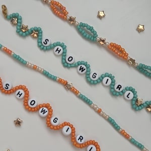 May include: Close-up of beaded bracelets in teal and orange, spelling out "SHOWGIRL" with white letter beads. Gold star-shaped beads and accents are interspersed throughout the design. The bracelets are on a white surface, with scattered gold star-shaped embellishments.