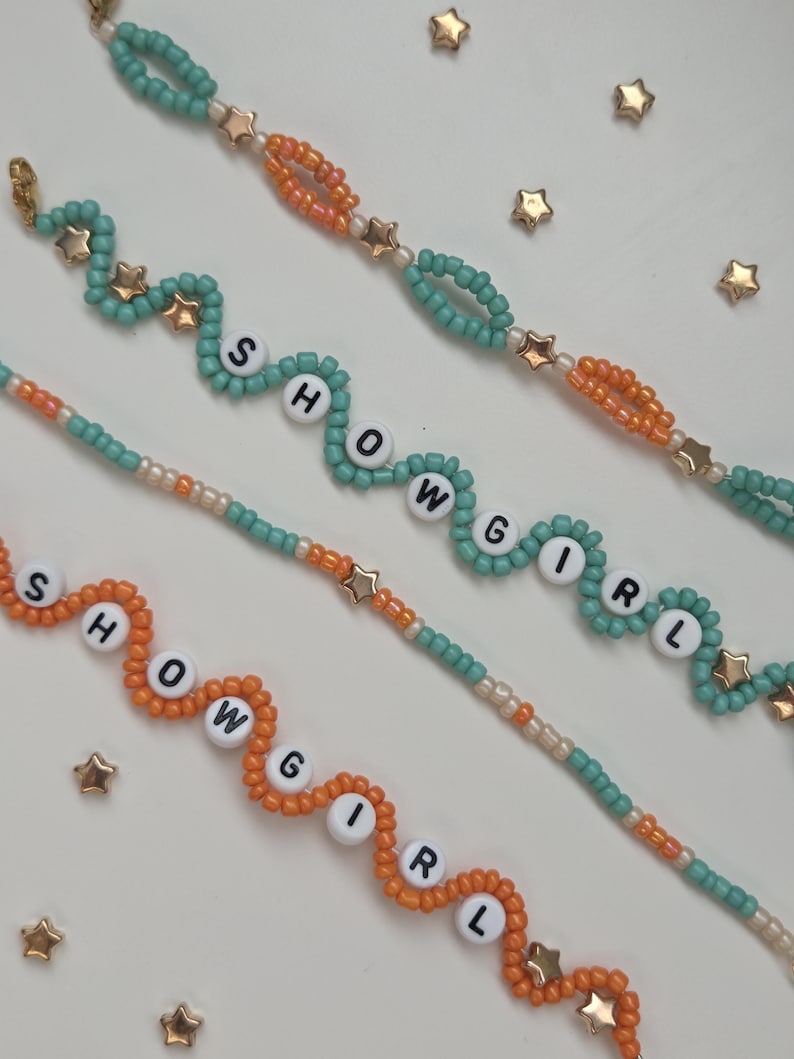 May include: Close-up of beaded bracelets in teal and orange, spelling out "SHOWGIRL" with white letter beads. Gold star-shaped beads and accents are interspersed throughout the design. The bracelets are on a white surface, with scattered gold star-shaped embellishments.