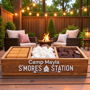 May include: A wooden s'mores station with compartments for biscuits, marshmallows, and chocolate. The station is labelled "Camp Mayla S'mores Station" in white lettering. The scene is set outdoors with a fire pit and seating.