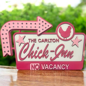 May include: A pink and white sign for "The Carlton Chick-Inn" with an arrow, stars, and a chicken graphic. The sign reads "No Vacancy" in pink letters. The sign is on a wooden surface.