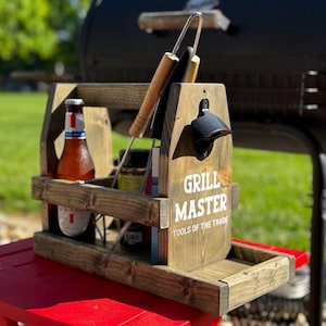 May include: A wooden grilling caddy with the words "Grill Master Tools of the Trade" printed on the side. The caddy holds grilling tools, condiments, and bottles of beer. An attached bottle opener is visible.