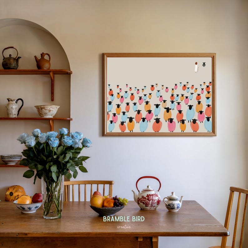 Op de afbeelding: Een ingelijste print van een kleurrijke illustratie van een kudde schapen. De schapen zijn in verschillende tinten roze, blauw en oranje. De print hangt aan een witte muur boven een houten tafel met een vaas met blauwe rozen en een theepot.