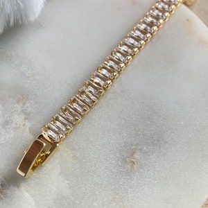 Puede incluir: Un brazalete de cadena dorada con piedras rectangulares transparentes. El brazalete est&aacute; sobre una superficie de m&aacute;rmol blanco.