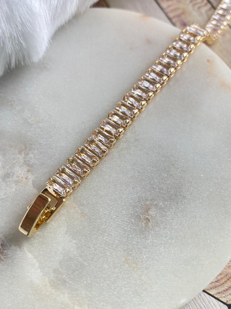 Puede incluir: Un brazalete de cadena dorada con piedras rectangulares transparentes. El brazalete est&aacute; sobre una superficie de m&aacute;rmol blanco.