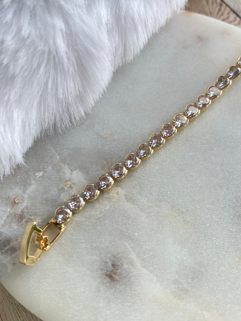 Puede incluir: Un brazalete de cadena dorada con piedras redondas transparentes. El brazalete tiene un cierre de broche.
