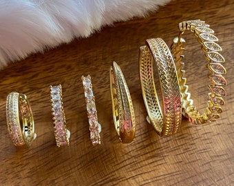 May include: A set of six gold hoop earrings with clear cubic zirconia stones. The earrings are of varying sizes and styles, including a pair with a delicate floral design.