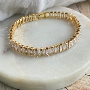 Puede incluir: Un brazalete dorado con una fila de piedras rectangulares transparentes. El brazalete est&aacute; sobre una superficie de m&aacute;rmol blanco.