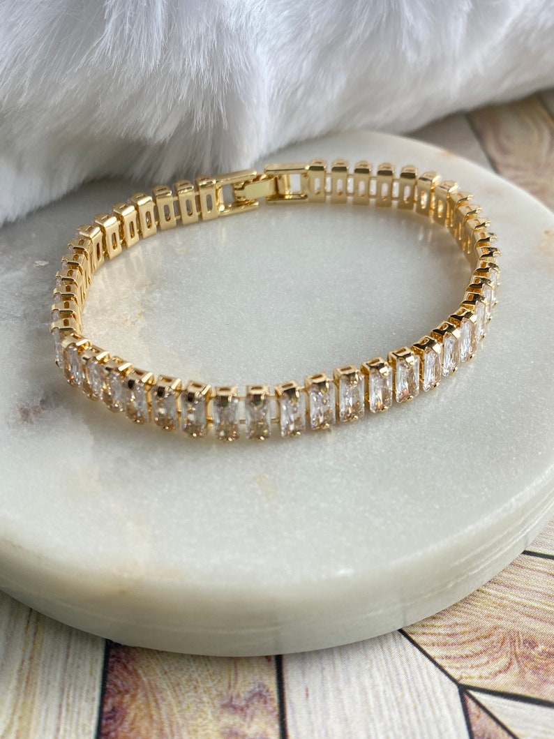 Puede incluir: Un brazalete dorado con una fila de piedras rectangulares transparentes. El brazalete est&aacute; sobre una superficie de m&aacute;rmol blanco.