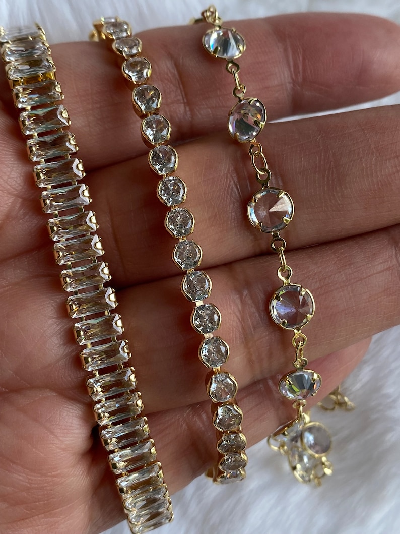 Puede incluir: Tres pulseras de oro con piedras de cristal transparentes. Las pulseras presentan diferentes estilos de piedras, incluyendo piedras de forma baguette y piedras redondas.