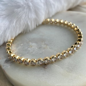 Puede incluir: Un brazalete dorado con una fila de piedras redondas transparentes.
