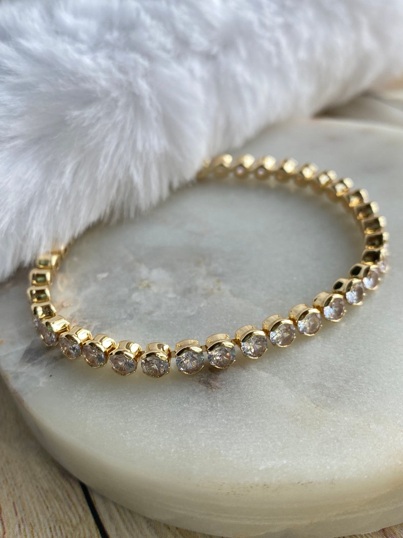 Puede incluir: Un brazalete dorado con una fila de piedras redondas transparentes.