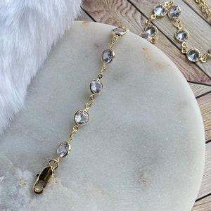 Puede incluir: Un brazalete de cadena dorada con piedras redondas transparentes. El brazalete tiene un cierre de langosta.