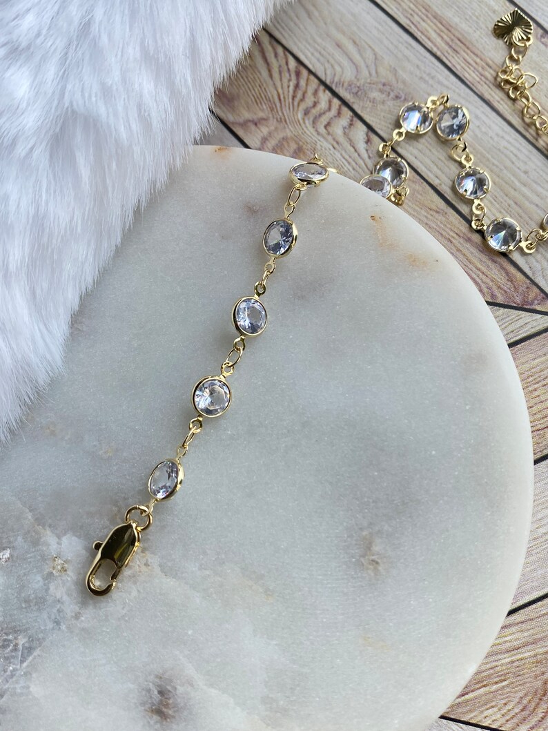 Puede incluir: Un brazalete de cadena dorada con piedras redondas transparentes. El brazalete tiene un cierre de langosta.