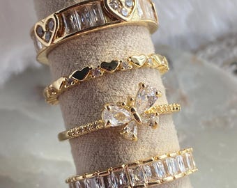 Puede incluir: Cuatro anillos dorados exhibidos sobre un fondo neutro. Los anillos presentan varios diseños, incluyendo detalles en forma de corazón, piedras con corte baguette y un motivo de mariposa. Los anillos probablemente son de metal chapado en oro y son adecuados para uso diario u ocasiones especiales.