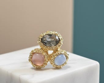 May include: A gold-toned ring featuring three gemstones: a large gray oval at the top, and smaller pink and blue ovals below. The gemstones are set in ornate, textured bezels.