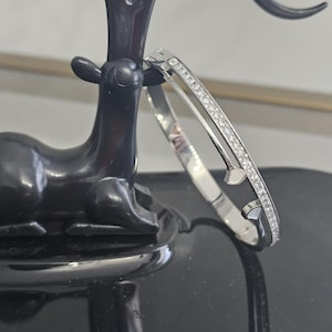May include: A silver-toned bracelet with a row of small, clear stones. The bracelet has a unique design with two curved sections and small, heart-shaped accents. The jewelry is displayed on a black decorative object.