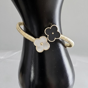 May include: Gold-tone bracelet with a textured band and clover-shaped accents. One clover is black, the other is white. The bracelet is displayed on a black mannequin arm against a neutral background. Jewelry.