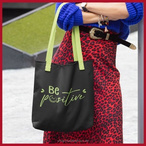 K&ouml;nnte beinhalten: Schwarze Tragetasche mit hellgr&uuml;nen Henkeln und dem Schriftzug "Be positive" in handschriftlicher Schrift. Die Tasche wird vor einem roten Leopardenmuster-Rock gehalten. Die Website shop.madebyfraus.de ist unten auf der Tasche aufgedruckt.