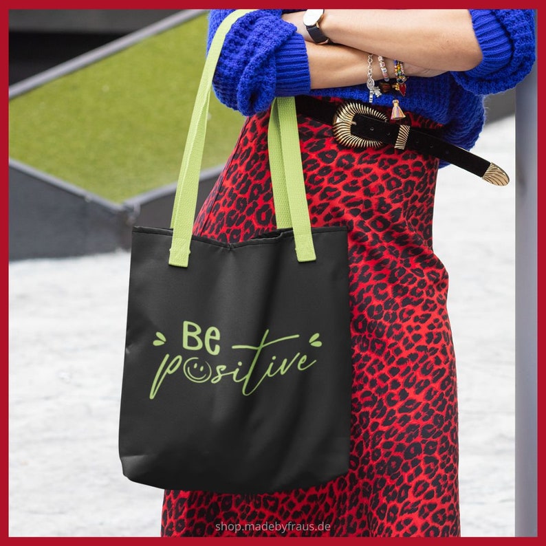 K&ouml;nnte beinhalten: Schwarze Tragetasche mit hellgr&uuml;nen Henkeln und dem Schriftzug "Be positive" in handschriftlicher Schrift. Die Tasche wird vor einem roten Leopardenmuster-Rock gehalten. Die Website shop.madebyfraus.de ist unten auf der Tasche aufgedruckt.