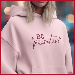 K&ouml;nnte beinhalten: Hellrosa Kapuzenpullover mit dem Schriftzug "Be positive" in dunkelrosa Schrift. Das Wort "positive" enth&auml;lt ein Smiley-Gesicht anstelle des Buchstabens "o". Der Hoodie hat eine Kapuze und eine Vordertasche.