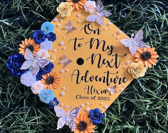May include: A yellow graduation cap decorated with paper flowers, butterflies, and pearls. The cap has the text "On to My Next Adventure" with the name "Alexia" and "Class of 2025". The flowers are in shades of blue, orange, and yellow.
