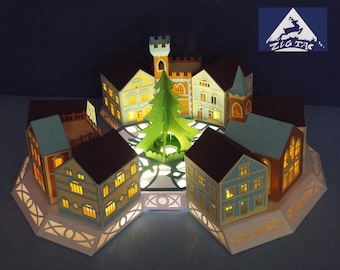 May include: A papercraft Christmas village scene featuring a green paper tree in the centre, surrounded by six houses with illuminated windows. The houses are painted in shades of blue, brown, and yellow. The base is white with a decorative pattern.