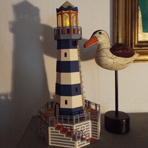 May include: A blue and white striped lighthouse with a yellow light in the top window. The lighthouse is on a wooden base with a brown and orange tiled floor. A white wooden seagull with a black beak is on a black stand to the right of the lighthouse.