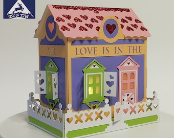 May include: A pink and purple papercraft house with a white picket fence. The house has a yellow roof with red hearts and the words "LOVE IS IN THE AIR" on the front.
