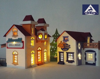May include: A miniature papercraft village scene with two buildings, a clock tower with a sign that reads "Jingle Bells" and a cafe with a sign that reads "Star Train Cafe". The buildings are lit with warm white lights and have a festive Christmas theme.