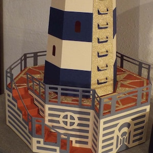 May include: A papercraft model of a lighthouse with a white and blue striped tower, a gold railing, and a red brick base. The lighthouse has a small window and a door.