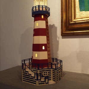May include: A papercraft lighthouse model with red and white stripes, a blue railing, and a gold glitter base. The lighthouse has a light inside and is lit up. The model is made by Zig Zag.
