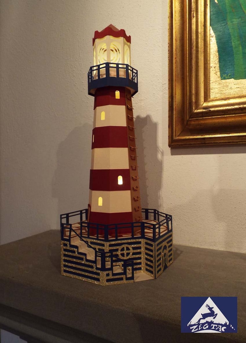 May include: A papercraft lighthouse model with red and white stripes, a blue railing, and a gold glitter base. The lighthouse has a light inside and is lit up. The model is made by Zig Zag.