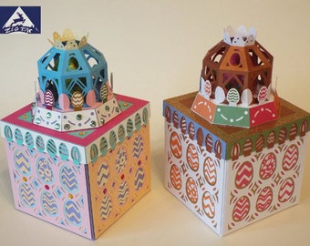 Könnte beinhalten: Zwei dekorative Osterboxen mit aufwendigen Designs. Die Boxen sind aus Papier und in verschiedenen Farben gehalten, darunter Rosa, Blau, Braun und Grün. Die Boxen sind mit einem kuppelförmigen Deckel versehen, der mit Eiern und einer Krone verziert ist.