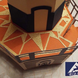 May include: A papercraft model of a lighthouse with a gold glitter-covered platform and a black railing. The lighthouse is made of brown and tan paper.