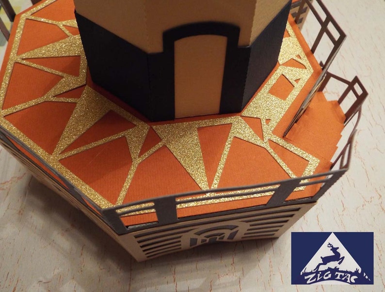 May include: A papercraft model of a lighthouse with a gold glitter-covered platform and a black railing. The lighthouse is made of brown and tan paper.