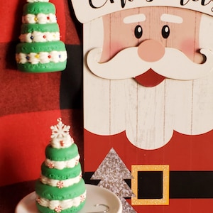 May include: Two green and white Christmas tree ornaments. One is on a white cake stand and the other is hanging from a red and black plaid fabric. The ornaments are decorated with white frosting, red and white candy canes, and green sprinkles. The background is a red and black plaid fabric with a wooden sign that says "Merry Christmas".