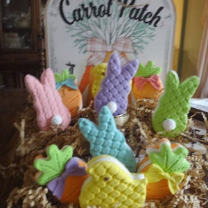 May include: A basket filled with pastel-colored bunny and chick-shaped cookies decorated with carrots and bows. The basket is sitting on a table with a sign that reads "Locally Grown Carrot Patch".