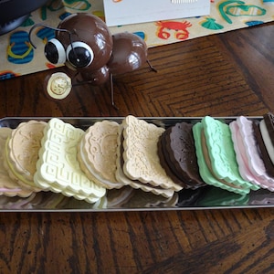 May include: A metal tray holds an assortment of colorful ice cream sandwiches. The sandwiches have various flavors, including chocolate, vanilla, and strawberry, with decorative patterns on the cookies. A whimsical brown ant figurine is in the background.