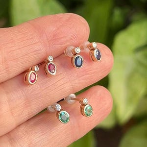 May include: Two pairs of stud earrings featuring oval-cut gemstones in gold settings. The top pair includes a red ruby and a dark blue sapphire, each accented with a small round diamond. The bottom pair features two green emeralds, each with a small round diamond accent.