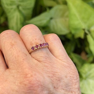 May include: A gold band ring featuring five small, round, vibrant red gemstones set in a row across the top. The metal band has a polished finish and the stones are held in place by small metal prongs, creating a continuous line of color.