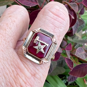 May include: A vintage gold-tone ring with a rectangular, deep red stone. The ring features a silver-tone emblem with a hammer, star, and other symbols. The ring is set against a background of green and burgundy foliage.