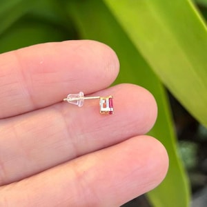 May include: A gold stud earring featuring a small, rectangular-cut pink gemstone set in a metal frame. The earring includes a clear plastic butterfly-style earring back attached to the thin gold post, showcasing a simple and functional design.