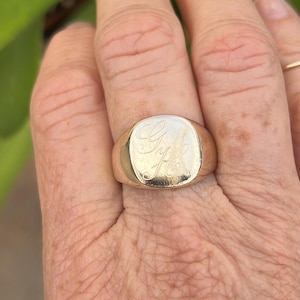 May include: A gold signet ring featuring a smooth, polished surface with an intricate, hand-engraved monogram of the letters G and H in a cursive script style. The ring has a rounded, solid band and a flat, oval-shaped face with a metallic finish.