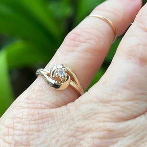 May include: A gold ring with a small, clear gemstone at the center. The ring has a unique, swirling design. The ring is on a finger, with a plain gold band visible on an adjacent finger. The background is a blurred green.