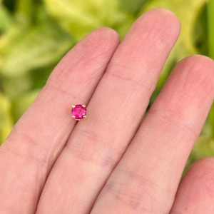 May include: A small, round, bright pink gemstone set in a gold metal setting. The gemstone is the focal point, showcasing its vibrant color and polished surface. The setting appears to have four prongs.