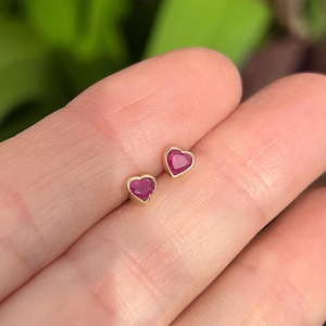 May include: A pair of heart-shaped stud earrings. Each earring features a vibrant red gemstone set in a gold-colored bezel. The earrings are small and delicate, perfect for everyday wear.